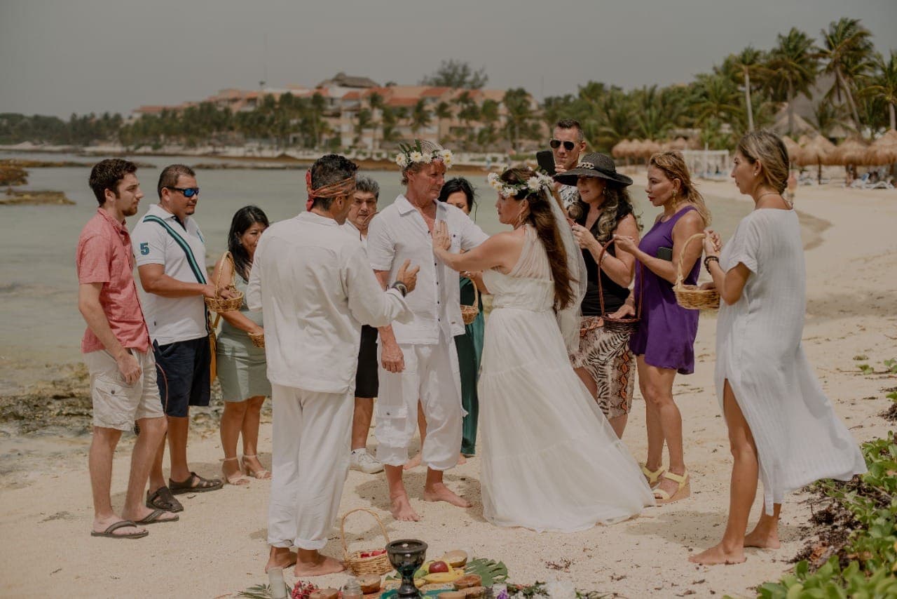 Pareja vestida de blanco celebra una ceremonia espiritual en la playa junto a sus invitados.