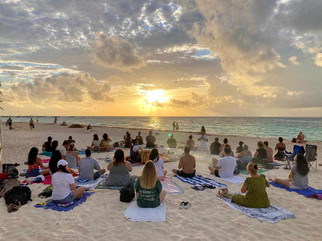 Grupo de personas meditando en la playa frente al mar durante un atardecer dorado.